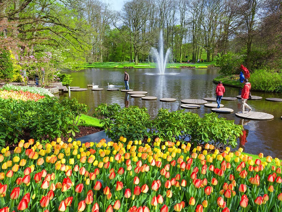 Lebendige rote und gelbe Tulpen im Vordergrund, Menschen gehen auf Trittsteinen über einen Teich in einem Frühlingsgarten.