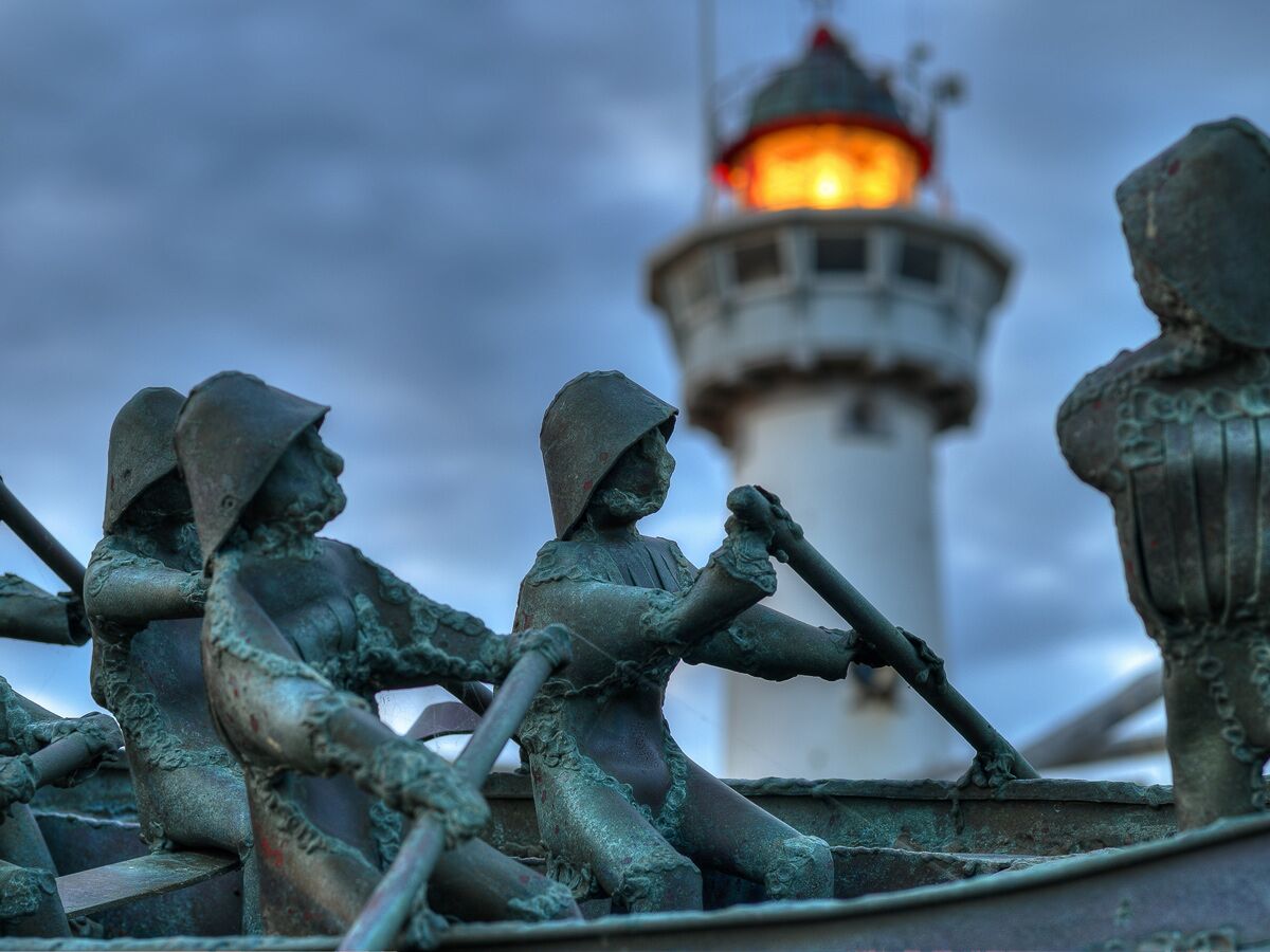 Detailaufnahme rudernder Bronze-Statuen in einem Boot. Im Hintergrund ein leuchtender, unscharfer Leuchtturm.