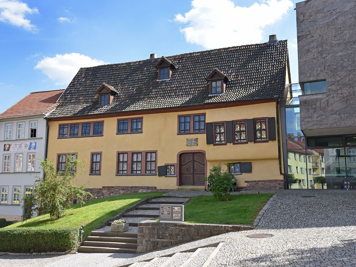 Gelbes Lutherhaus Mansfeld mit modernem Anbau, umgeben von Grünflächen und Kopfsteinpflaster, unter blauem Himmel.