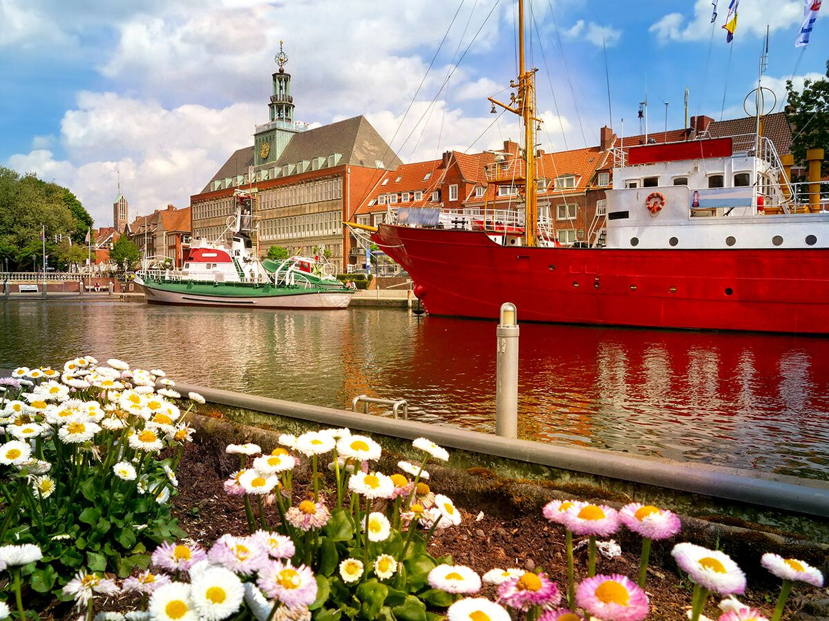 Historisches Rathaus Leer am Hafen mit roten und grünen Booten, blühenden Margeriten im Vordergrund an einem sonnigen Tag.