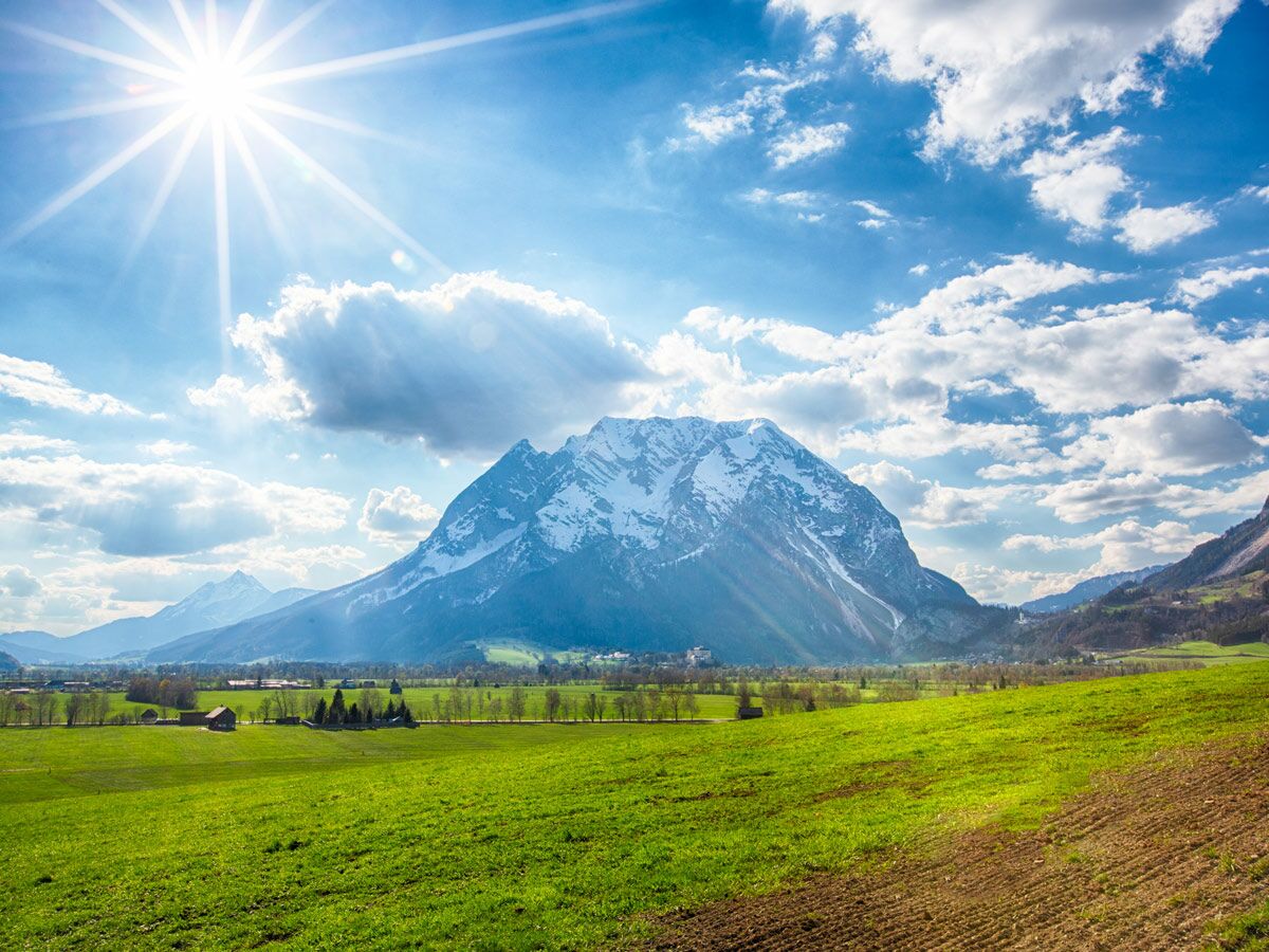 Sonnenbeschienene Alpenlandschaft mit schneebedecktem Berg, grünen Wiesen und blauem Himmel mit weißen Wolken.
