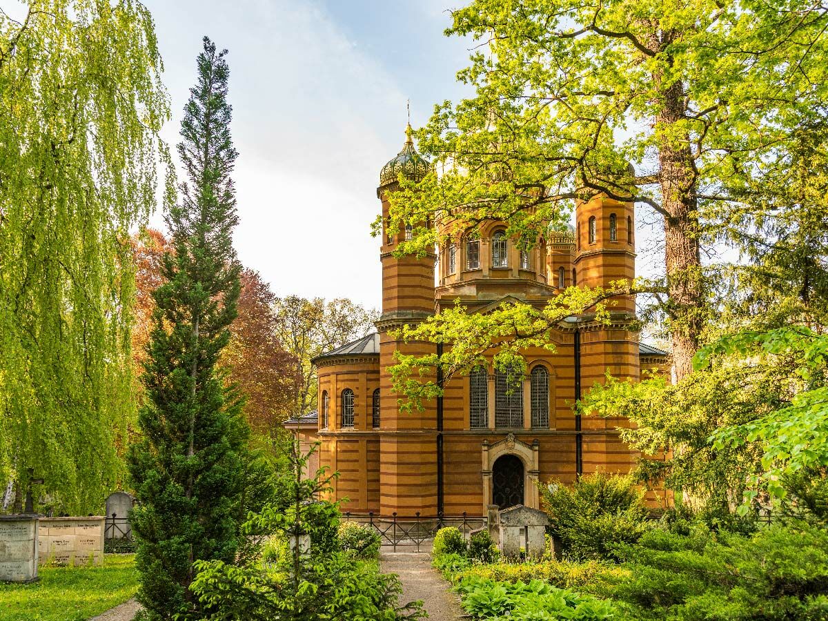 Eine orangerote Backsteinkirche mit Kuppeln und Türmen, umgeben von üppig grünen Bäumen und Büschen in einem Friedhof oder Park.