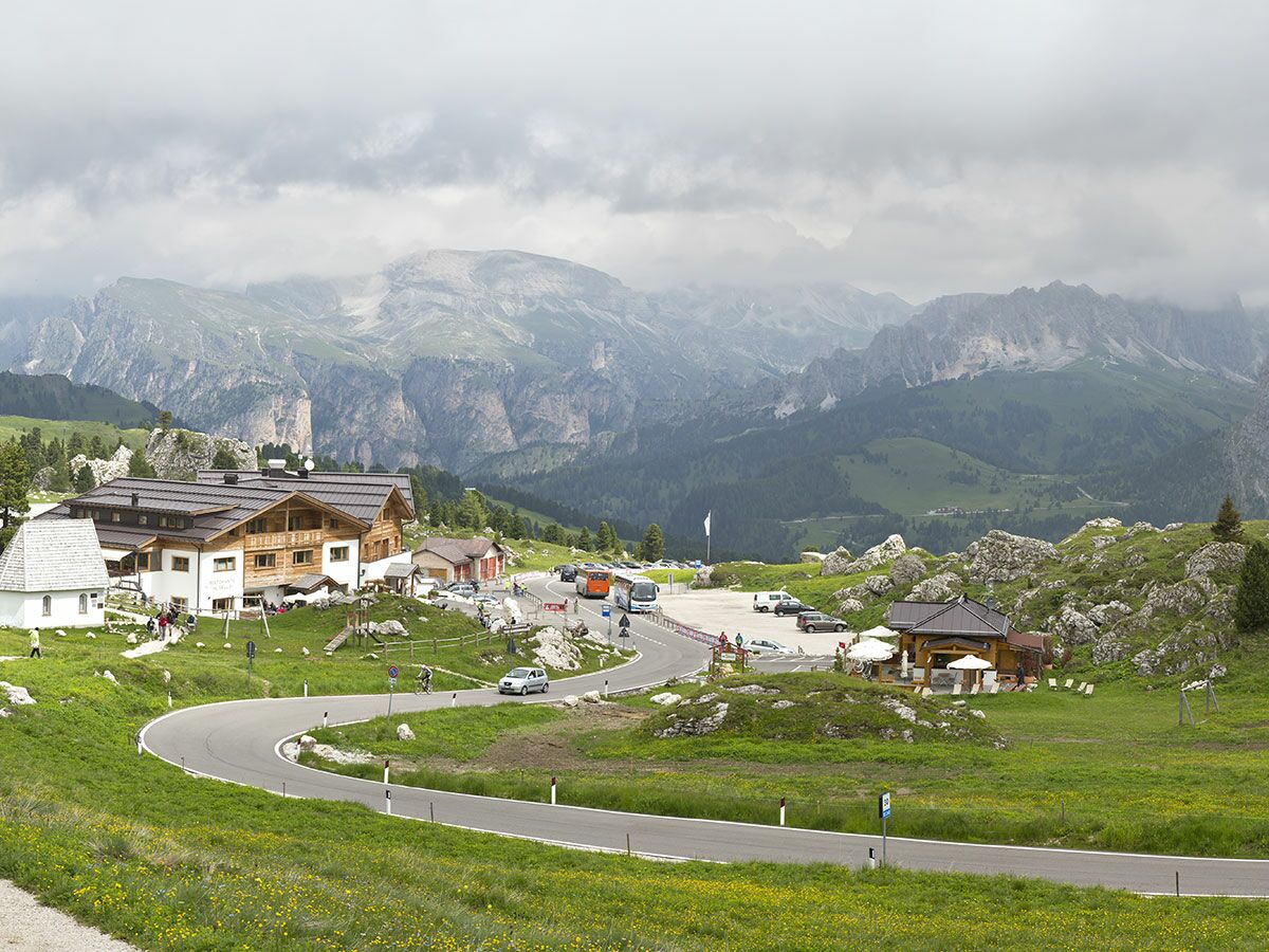 Serpentinenstraße im Alpenpass mit Berghütte, Restaurant und Autos unter bewölktem Himmel. Grüne Berglandschaft, Dolomiten.