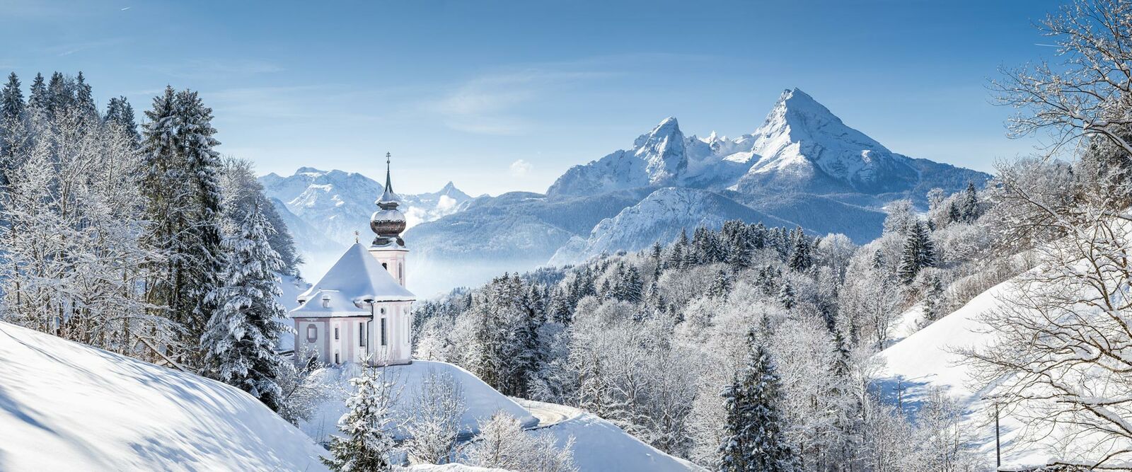Idyllische Kirche St. Sebastian in Ramsau, umgeben von verschneiten Bäumen und dem Watzmann-Massiv unter blauem Himmel.