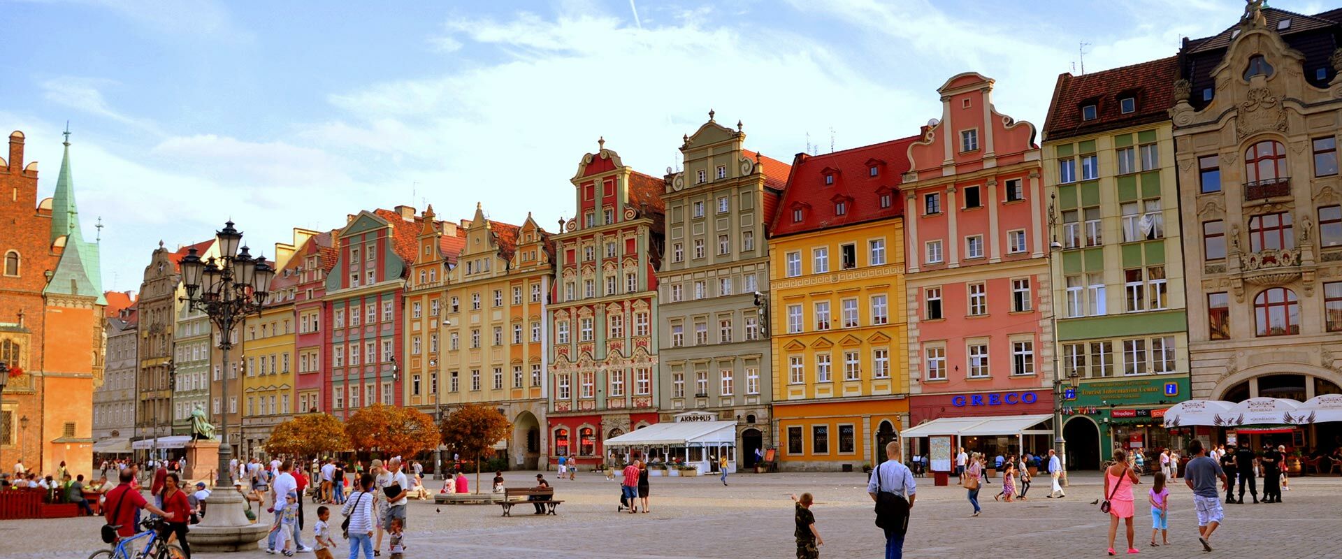 Bunter Marktplatz von Wrocław, Polen, mit historischen Gebäuden, Menschen und einem Kirchturm unter blauem Himmel.