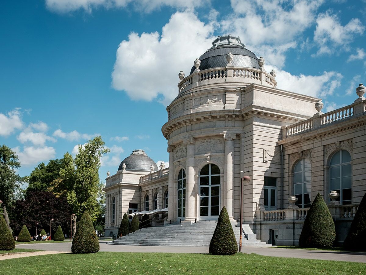 Großes, helles Palastgebäude mit runden Kuppeln und aufwendiger Steinfassade an einem sonnigen Tag im Park.