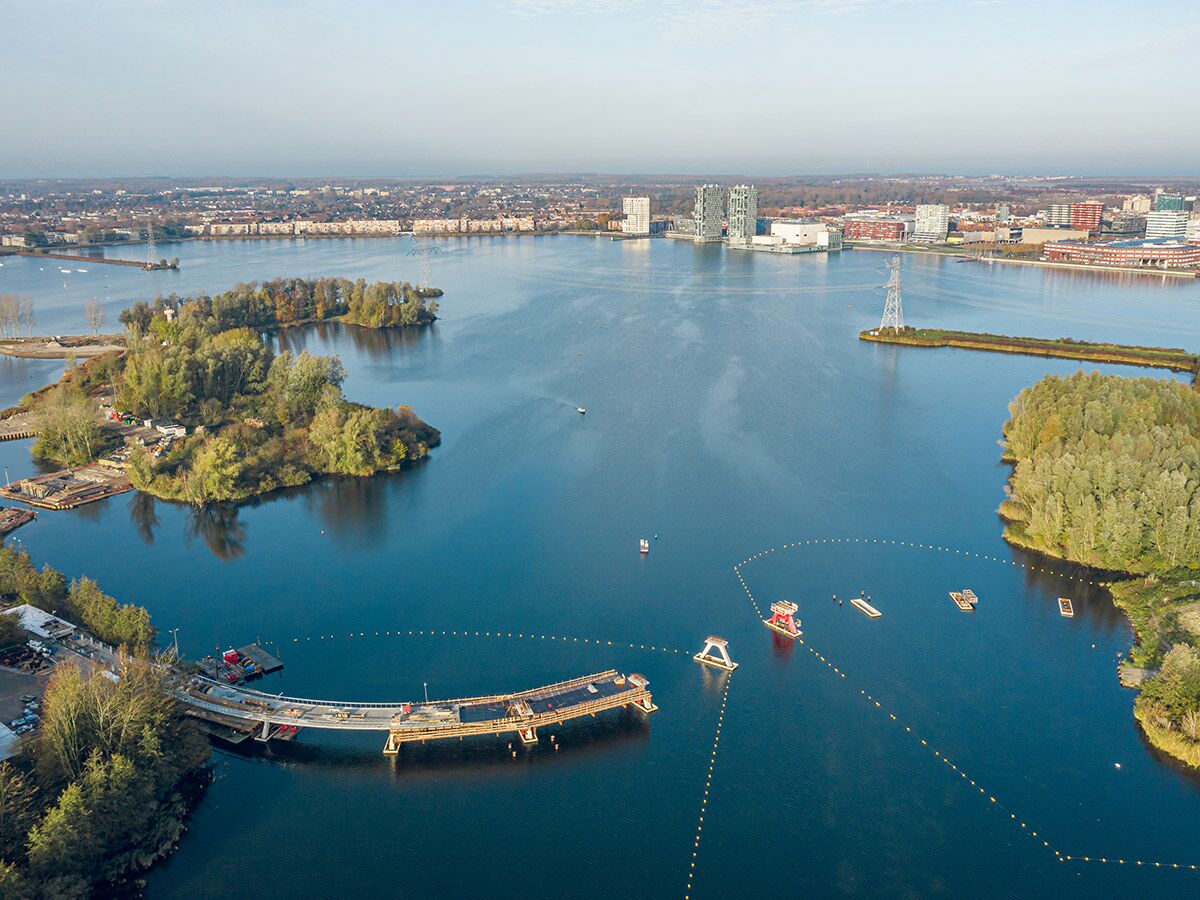Luftaufnahme eines großen Sees mit einer geschwungenen Brücke im Bau, bewaldeten Inseln und einer modernen Stadt im Hintergrund.