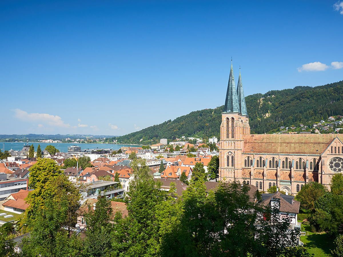 Panoramablick auf das Konstanzer Münster, die Stadt Konstanz am Bodensee und bewaldete Hügel unter blauem Himmel.