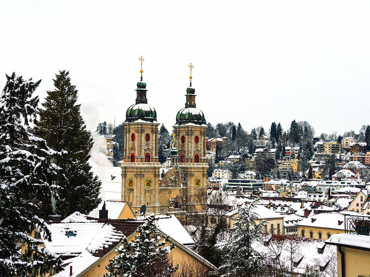Winterliche Stadtansicht von St. Gallen mit der verschneiten Stiftskirche und Dächern unter weißem Himmel.