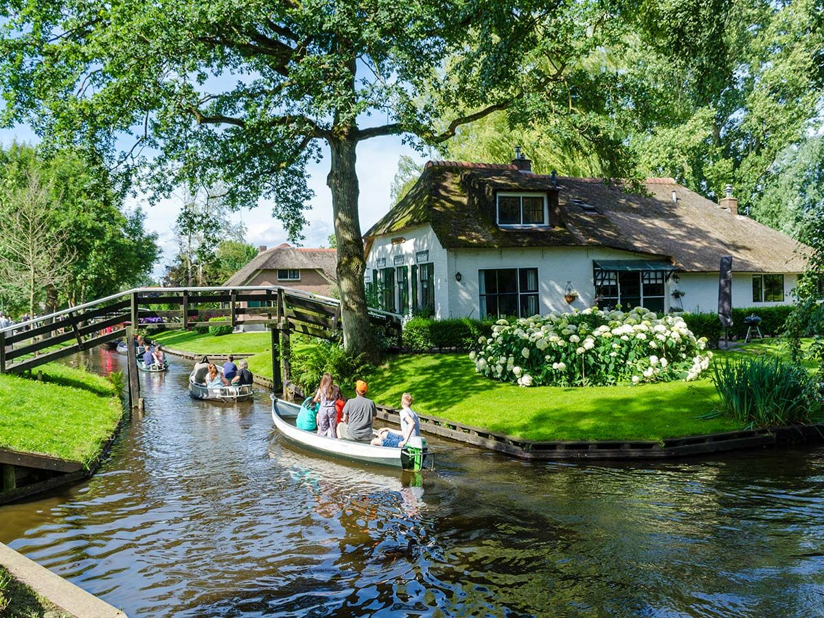 Menschen fahren in Booten auf einem Kanal durch das idyllische Dorf Giethoorn mit reetgedeckten Häusern und grüner Natur.