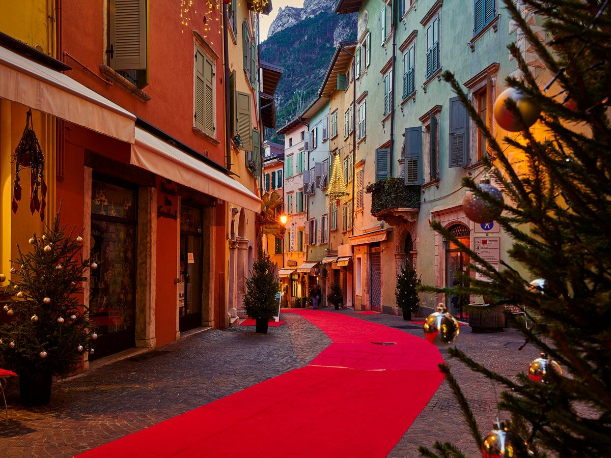 Festlich dekorierte Altstadtgasse mit rotem Teppich, Weihnachtsbäumen, Lichtern und bunten Häusern vor Bergkulisse.