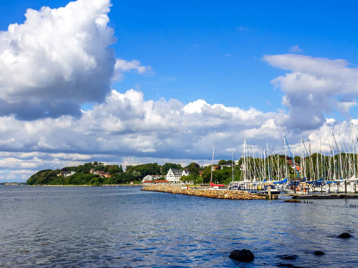 Blick auf einen Hafen mit vielen Segelbooten am Steg, umgeben von grüner Küstenlandschaft und Häusern unter blauem Himmel mit weißen Wolken.