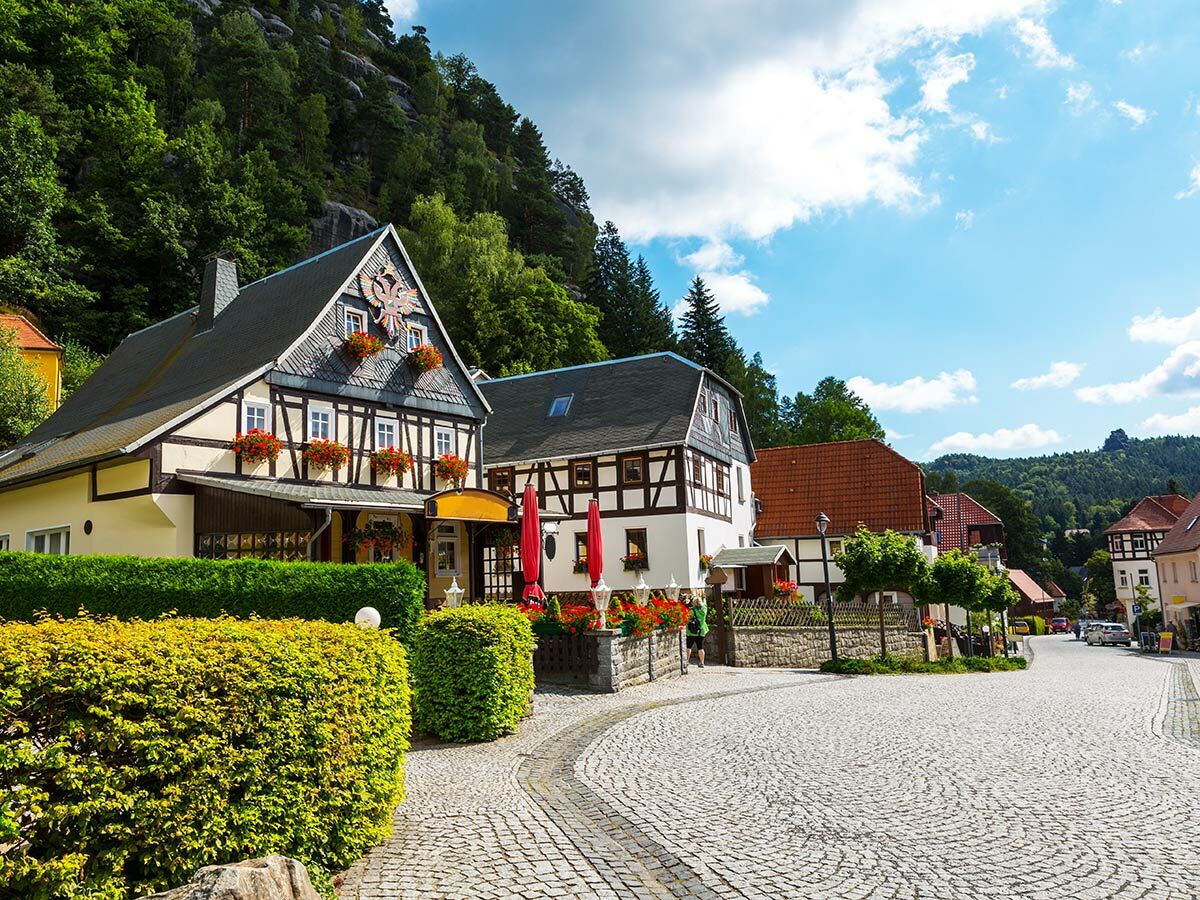 Idyllisches deutsches Dorf mit Fachwerkhäusern, Blumenkästen und Kopfsteinpflasterstraße vor bewälderten Hügeln.