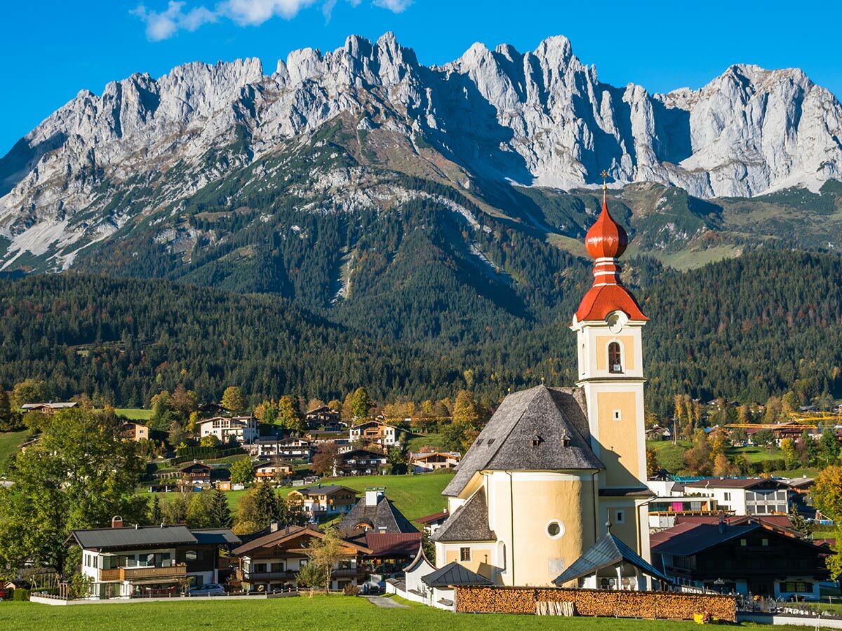 Eine malerische Alpenkirche mit rotem Zwiebelturm in einem Bergdorf vor majestätischen Felsbergen und grünen Wäldern im Herbst.
