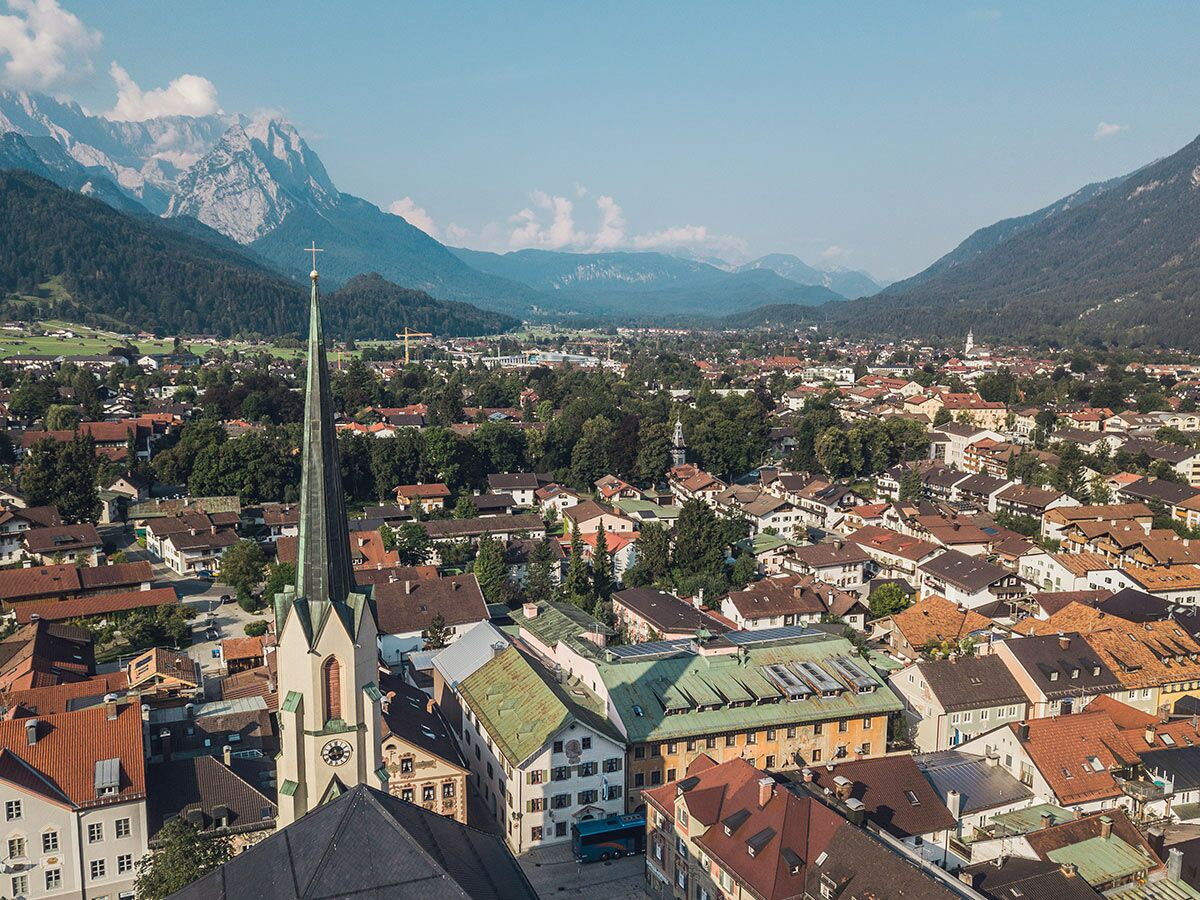 Luftaufnahme von Garmisch-Partenkirchen mit Kirchturm, Dächern und Wettersteinbergen unter blauem Himmel.