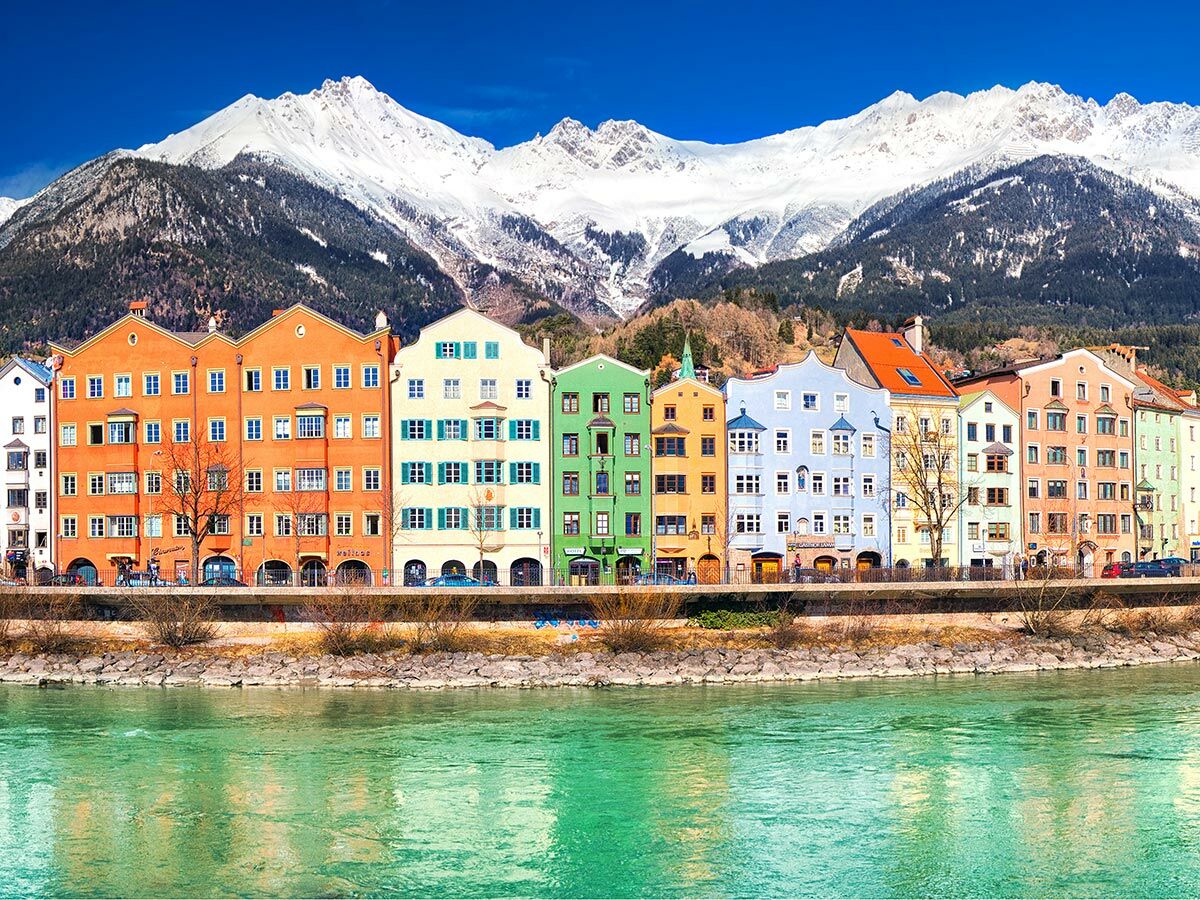 Bunte Häuserreihe am Inn-Fluss in Innsbruck mit schneebedeckten Alpen im Hintergrund bei strahlendem Sonnenschein.