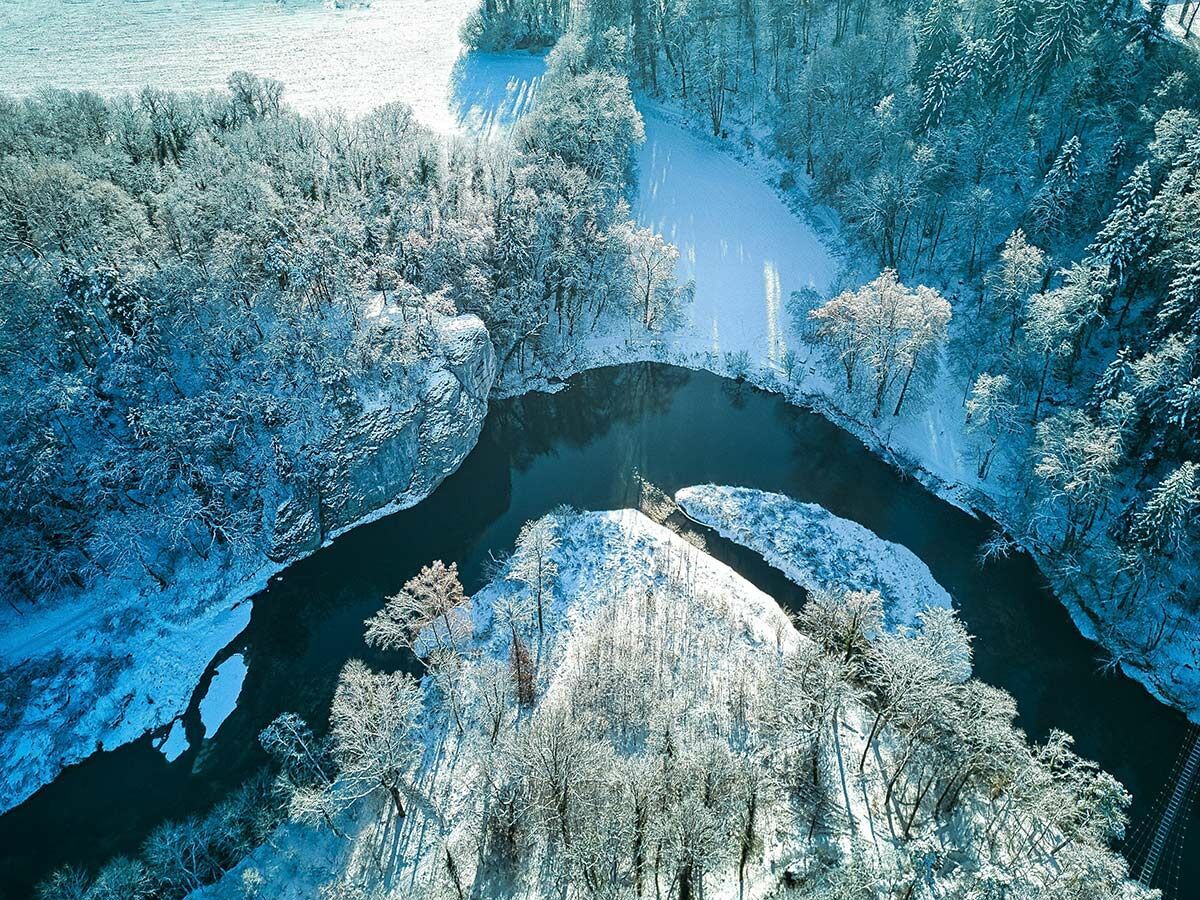 Luftaufnahme eines gewundenen Flusses durch eine schneebedeckte Winterlandschaft mit Felsen und Bäumen im Sonnenlicht.