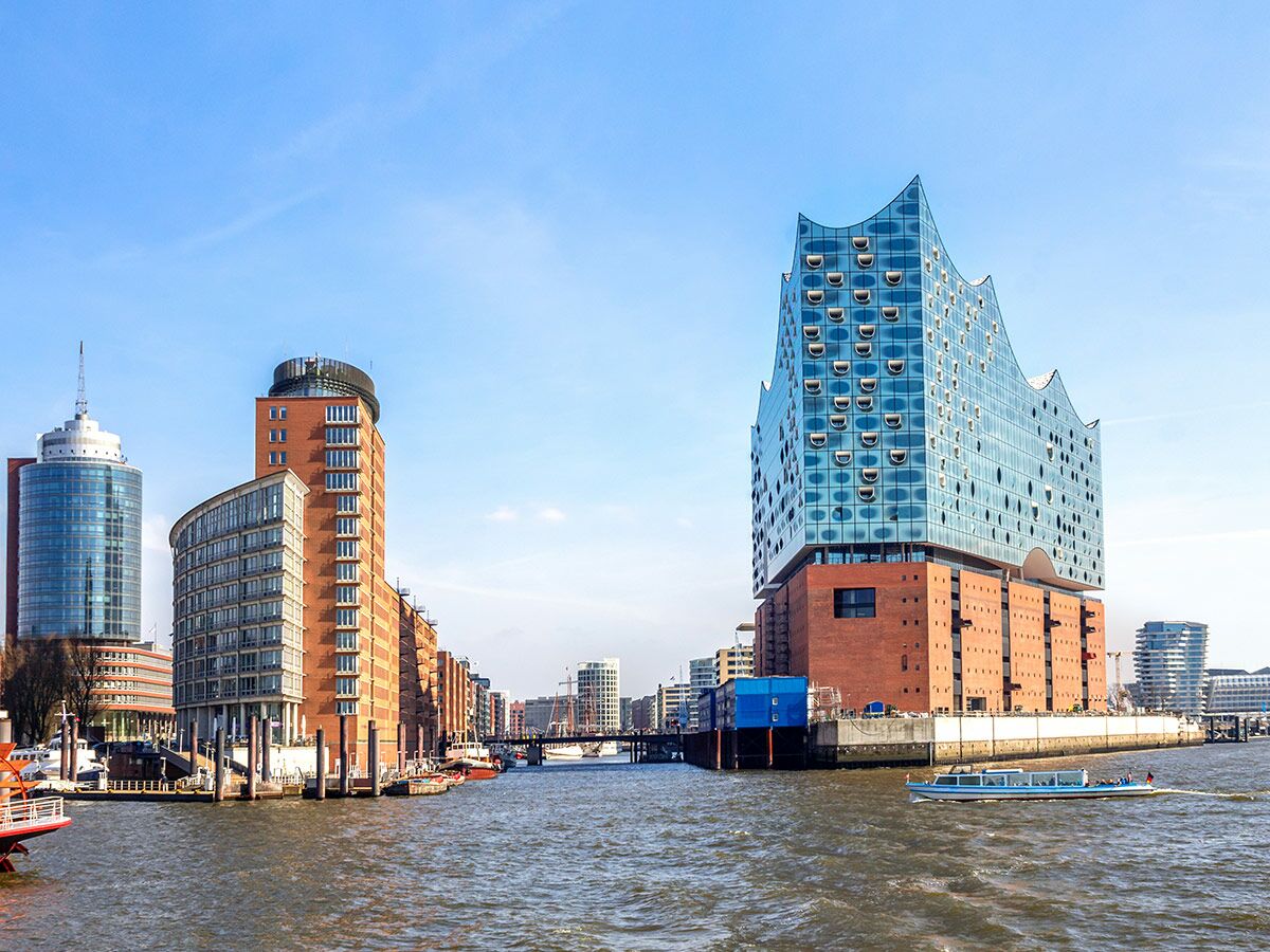 Die Elbphilharmonie in Hamburg am Wasser, umgeben von modernen Gebäuden und Booten im Hafen unter blauem Himmel.