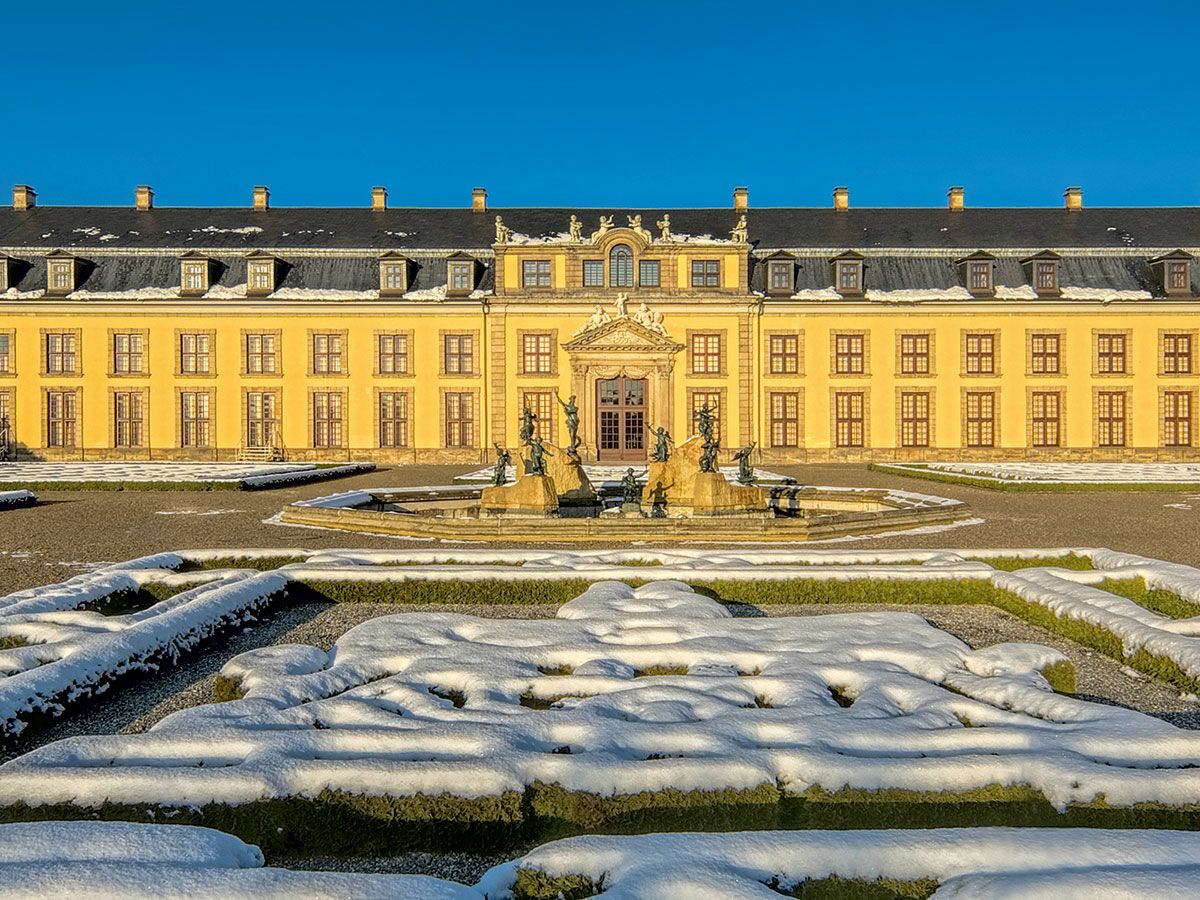 Prächtiger gelber Palast mit Barockgarten, schneebedeckten Hecken und Brunnen unter blauem Winterhimmel.