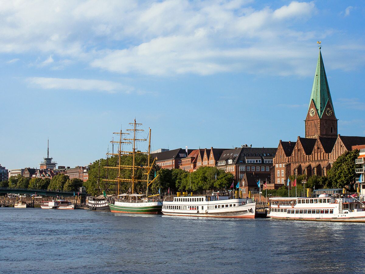 Blick auf Bremens Schlachte Promenade an der Weser mit der Martinikirche, dem Segelschiff Alexander von Humboldt und Fahrgastschiffen bei sonnigem Wetter.