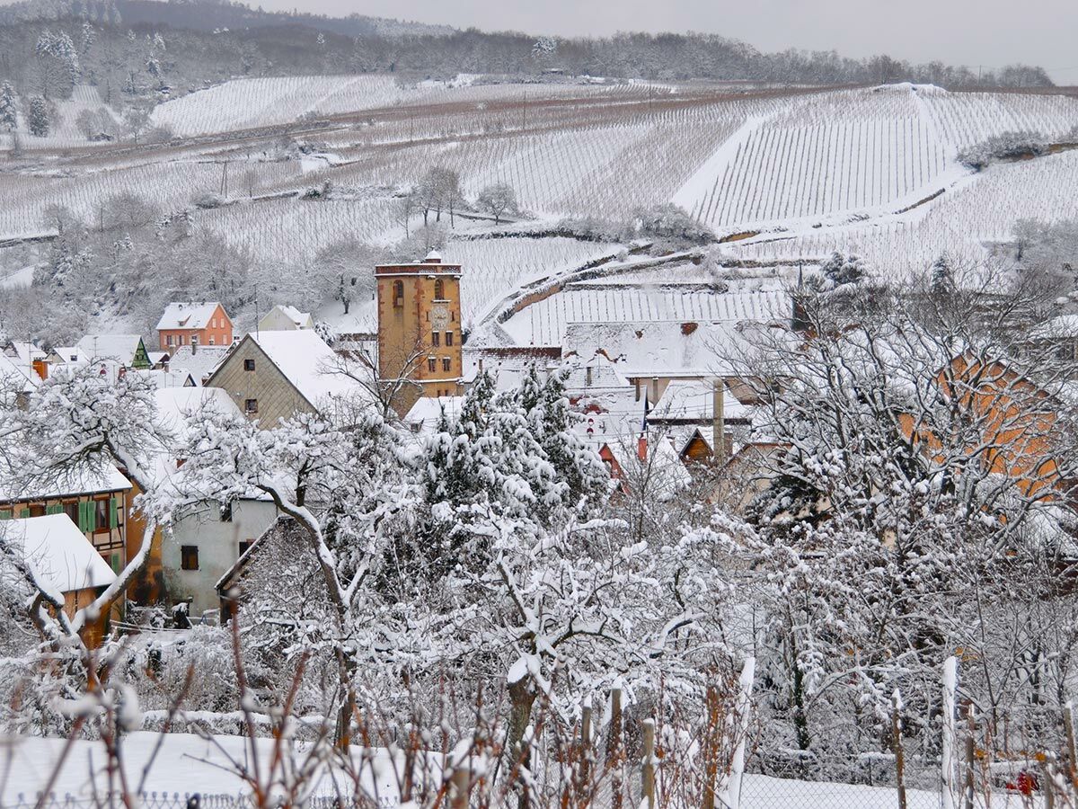 Verschneites Dorf im Winter mit prominentem Kirchturm, traditionellen Häusern und Weinbergen auf den umliegenden Hügeln.