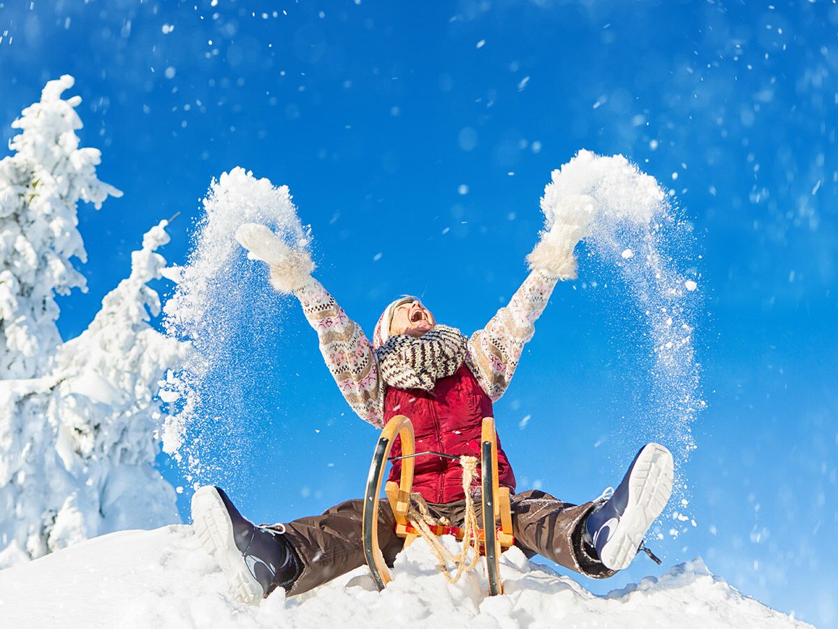 Lachende Frau auf Schlitten wirft Schnee in die Luft unter blauem Himmel, umgeben von verschneiten Bäumen. Winterfreude.