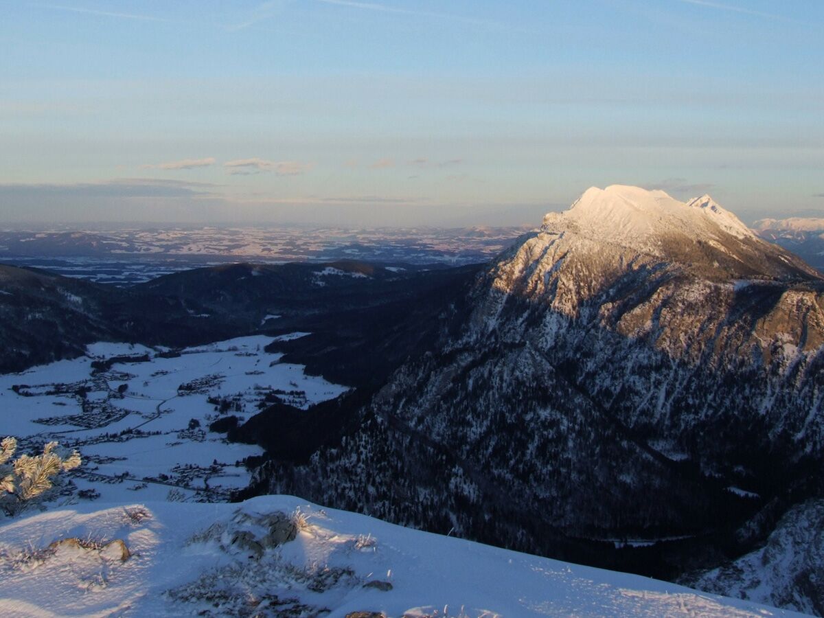 Panoramablick auf eine winterliche Alpenlandschaft mit schneebedeckten Gipfeln, sonnenbeschienenem Tal und Dorf. Blauer Himmel, weite verschneite Ebenen.
