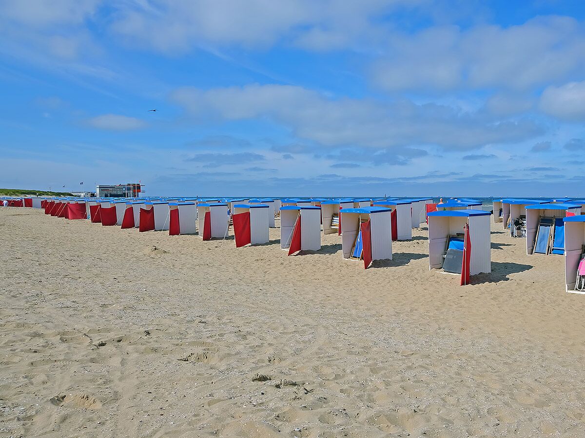 Rote und weiße Strandhütten auf sonnigem Sandstrand mit Meer und blauem Himmel im Hintergrund, wenige Personen.