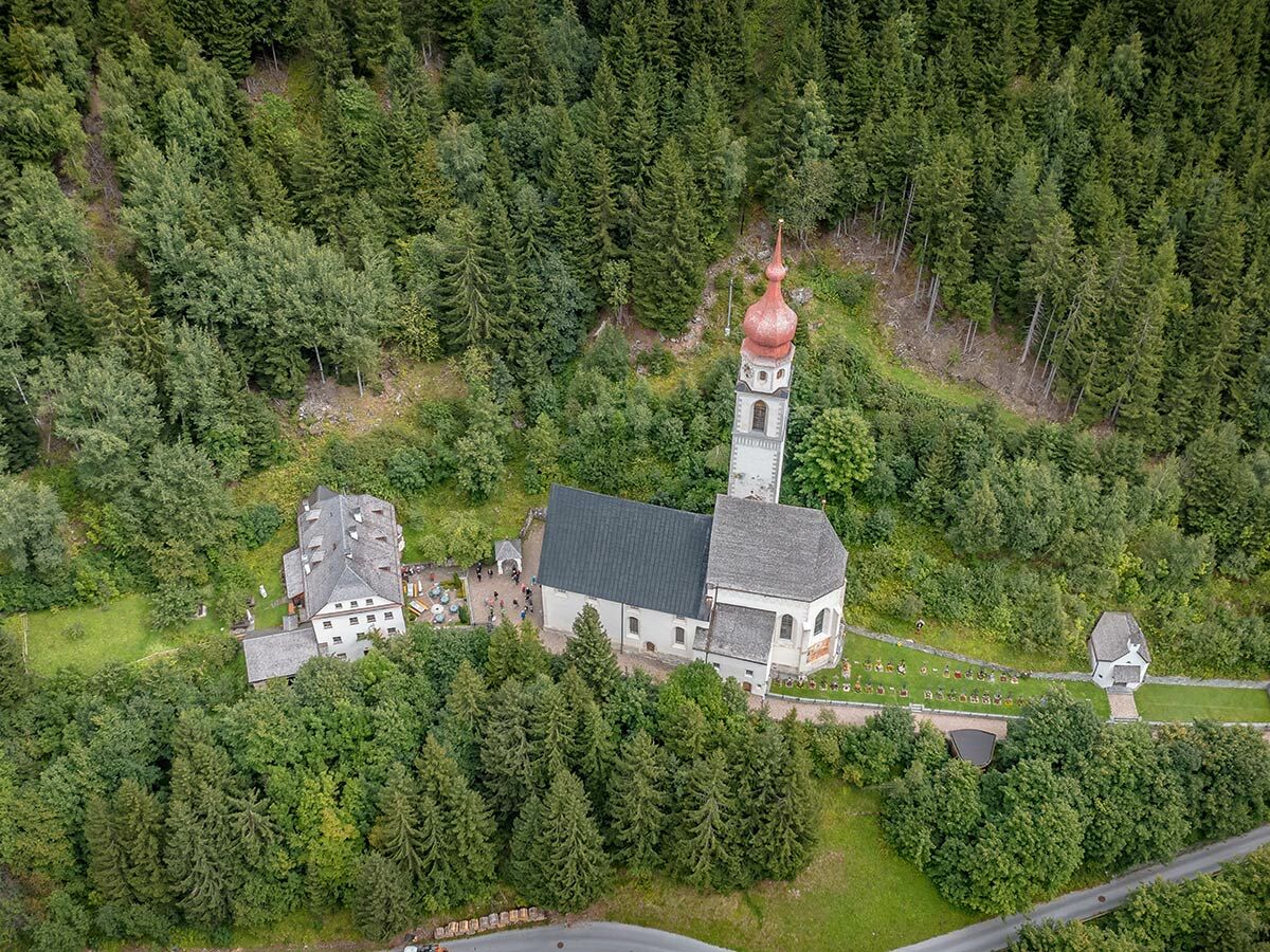 Luftaufnahme einer historischen Kirche mit rotem Zwiebelturm, Friedhof und weiteren Gebäuden im dichten Bergwald.