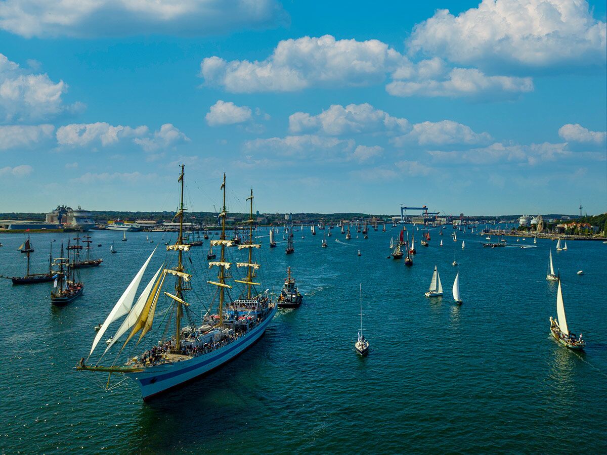 Großsegler mit weißen Segeln und vielen Menschen auf dem Wasser während einer Parade mit zahlreichen Segelbooten und Schiffen, Hafenstadt im Hintergrund.