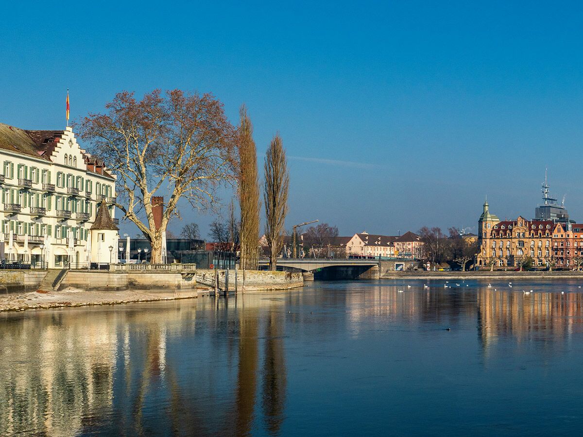 Winterlicher Blick auf Konstanz mit Steigenberger Inselhotel, Konzilgebäude, Rhein und Brücke unter blauem Himmel mit Wasservögeln.