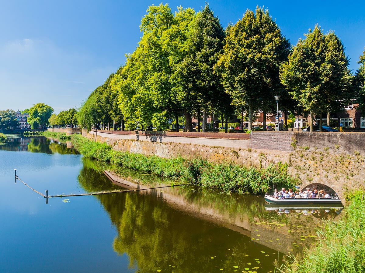 Bootsfahrt auf einem sonnigen Stadtkanal mit grünen Bäumen, alter Steinmauer und Menschen, die die Tour genießen.