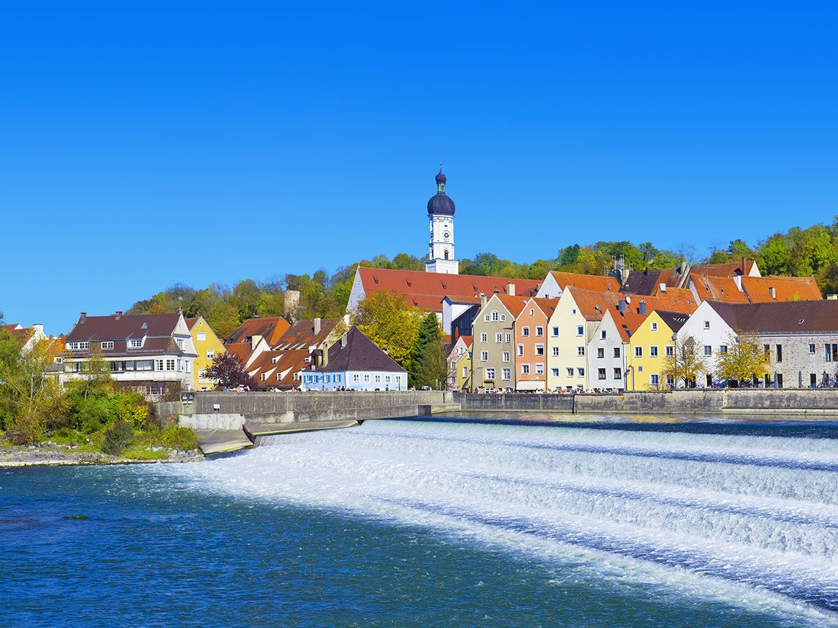 Malerische Altstadt Landsberg am Lech mit historischen, bunten Häusern am Fluss und weitem Wasserfallwehr unter blauem Himmel.