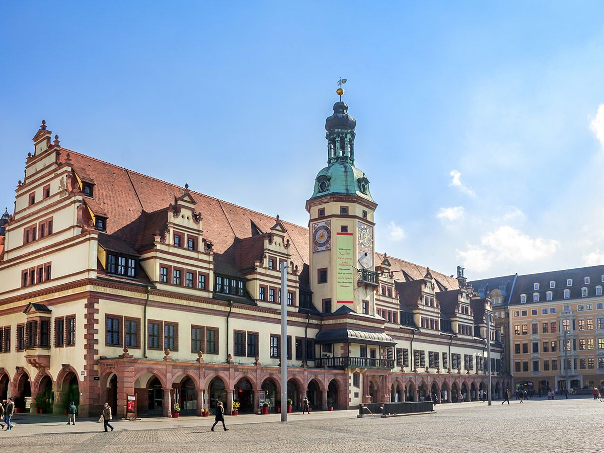 Historisches Altes Rathaus in Leipzig mit Turm, Uhr, rotem Dach, Arkaden und Menschen auf dem Marktplatz unter blauem Himmel.