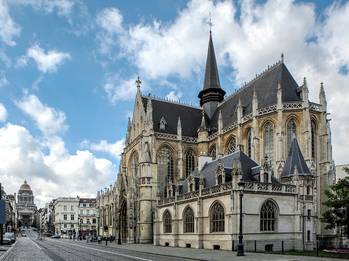Ansicht der imposanten gotischen Kirche und einer belebten Straße in Brüssel unter blauem Himmel mit Wolken. Im Hintergrund der Justizpalast.