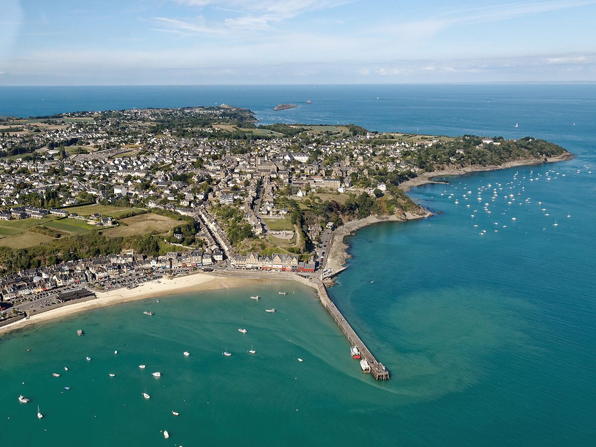 Luftbild einer lebhaften Küstenstadt in der Bretagne mit Sandstrand, einem langen Pier und vielen Booten im türkisblauen Meer an einem sonnigen Tag.