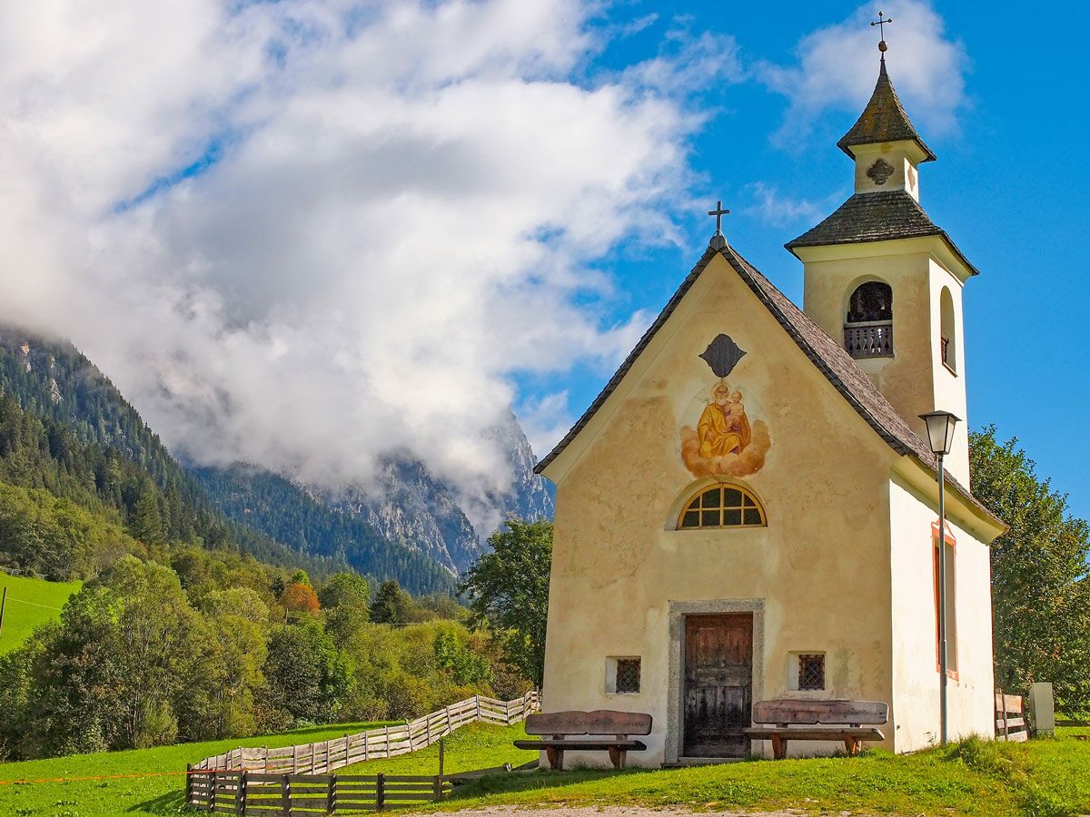 Malerische Bergkapelle mit Glockenturm und Fresko, umgeben von grünen Wiesen und bewölkten Alpenbergen.