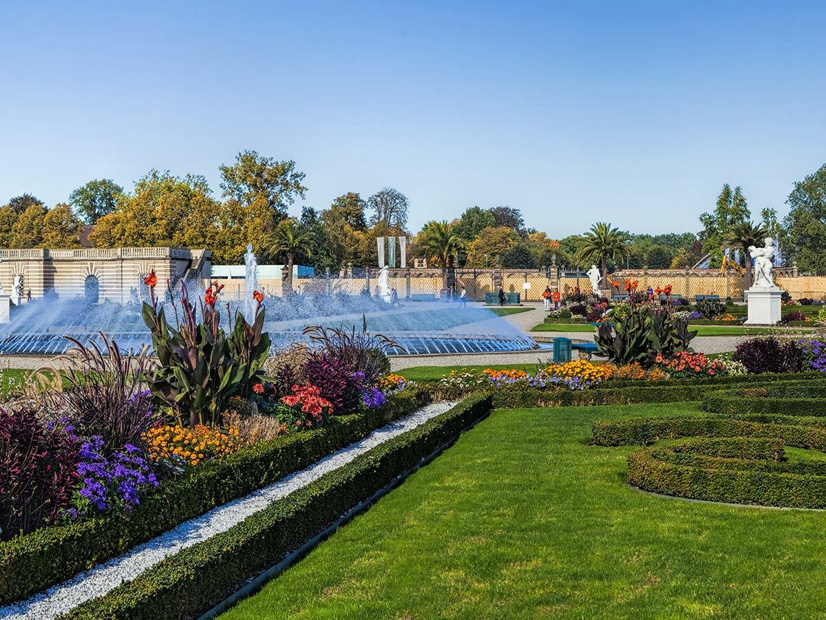 Prächtiger Barockgarten mit großer Fontäne, bunten Blumenbeeten, grünen Hecken, weißen Statuen und klassischer Architektur.