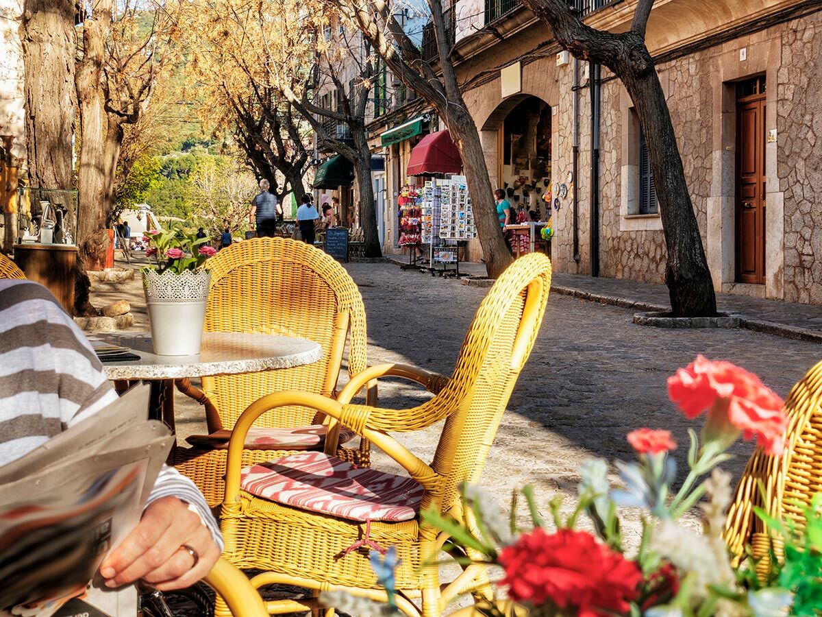 Entspannter Blick auf ein sonniges Straßencafé mit Rattanmöbeln und Blumen, während eine Person Zeitung liest.