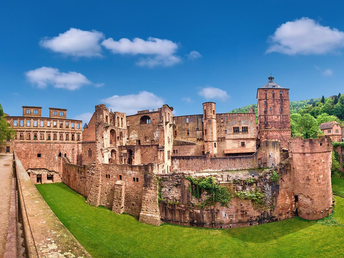 Blick auf die imposante Ruine des Heidelberger Schlosses mit roten Sandsteinmauern, grünen Wiesen und blauem Himmel.