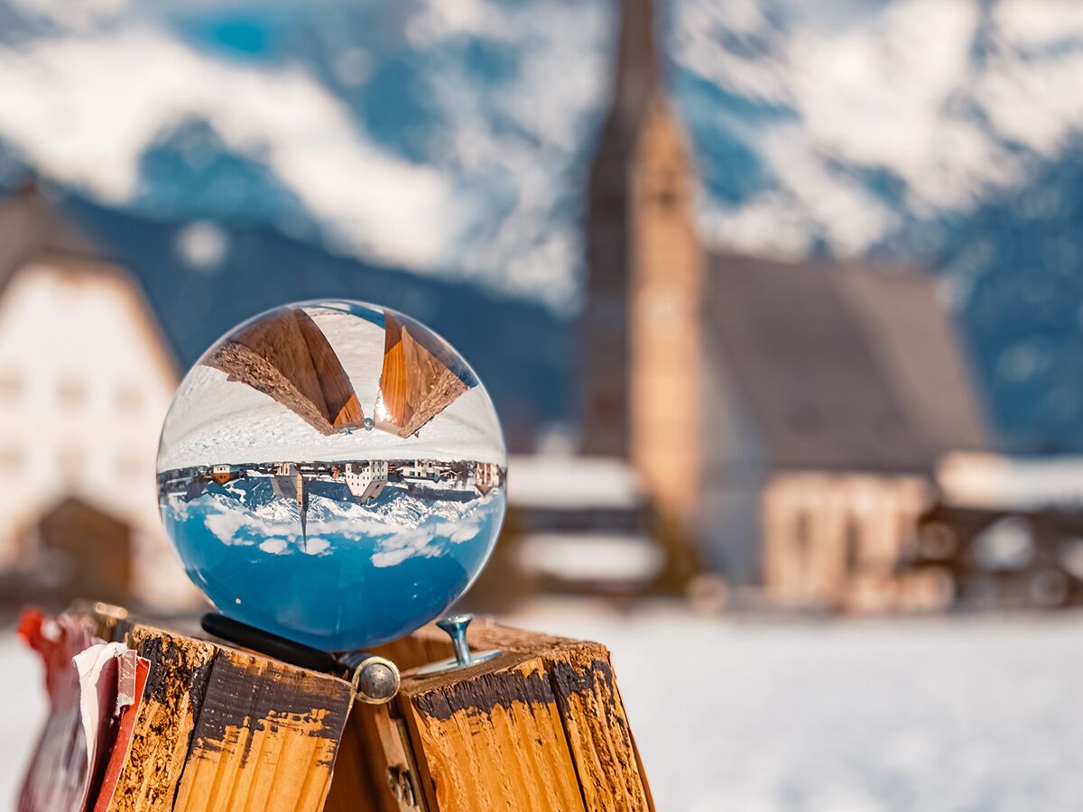 Eine Kristallkugel spiegelt eine verschneite Winterlandschaft mit Kirche, Häusern und Bergen auf einem Holzbalken.