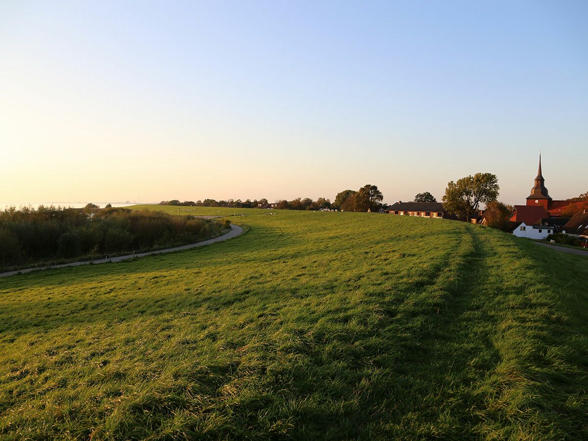 Weitblick über eine grüne Deichlandschaft mit gewundenem Weg, Häusern und Kirchturm im goldenen Licht des Sonnenuntergangs.