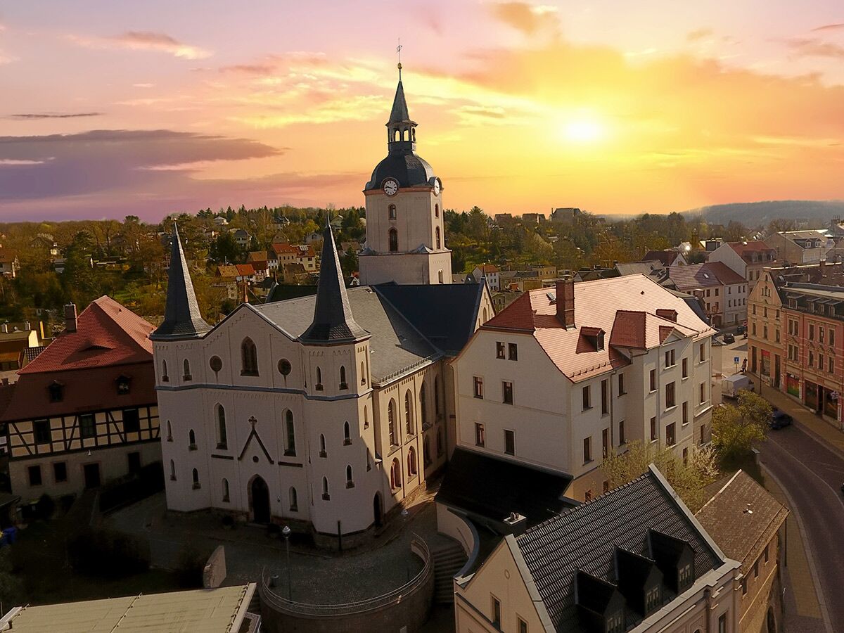 Historische Kirche mit Glockenturm und spitzen Türmen in einer europäischen Stadt bei goldenem Sonnenuntergang, umgeben von Gebäuden und Straßen.