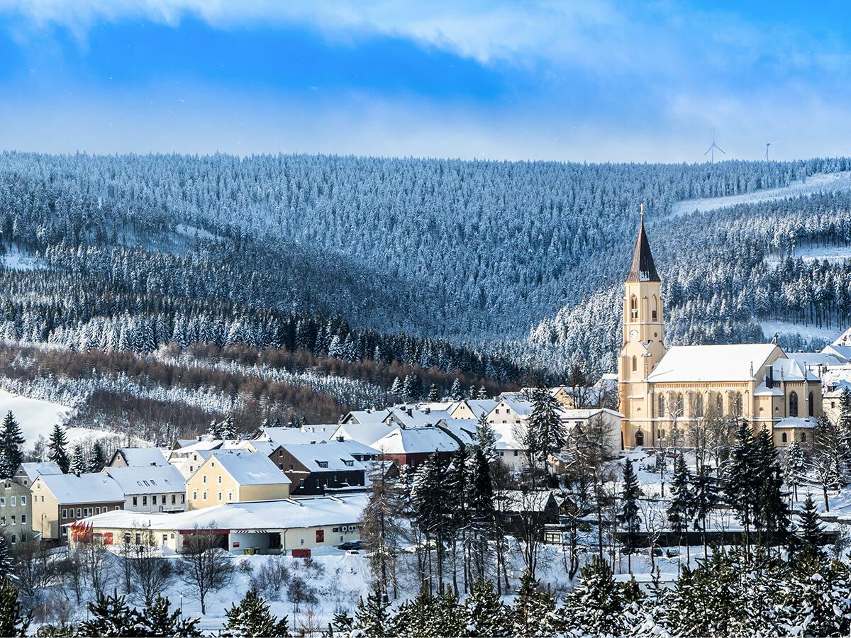 Verschneites Dorf mit großer Kirche, umgeben von schneebedeckten Nadelwäldern unter blauem Himmel, Windräder am Horizont.