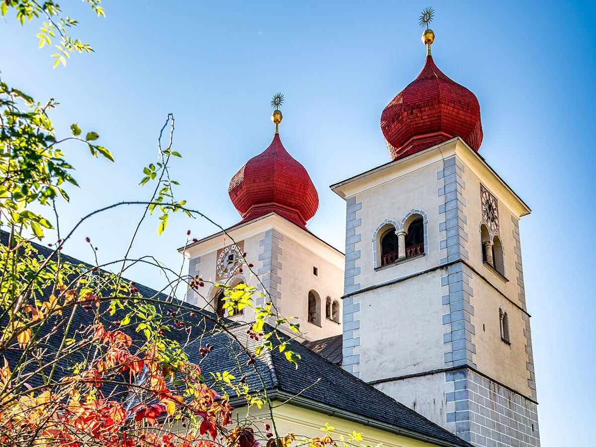 Zwei barocke Kirchtürme mit roten Zwiebelhauben unter blauem Himmel, umrahmt von herbstlichem Laub und Beeren.