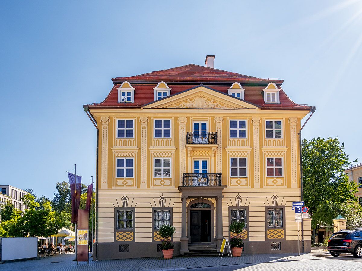 Prächtiges barockes Stadthaus mit gelber Fassade, rotem Dach, verzierten Fenstern und Balkonen unter blauem Himmel in einer Stadt.
