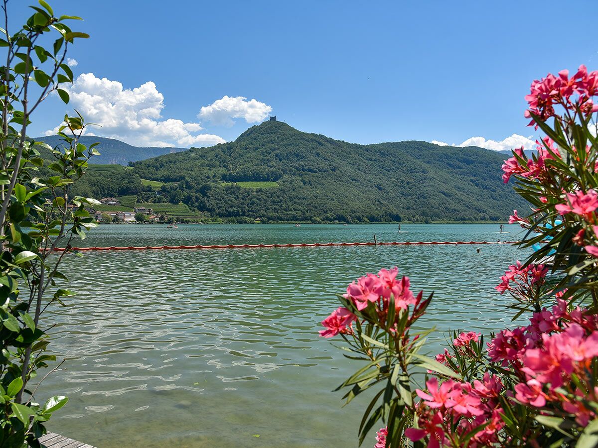 Idyllischer See mit grünen Bergen, historischem Schloss auf Gipfel und rosa Oleanderblüten im Vordergrund; Menschen paddeln auf dem Wasser.