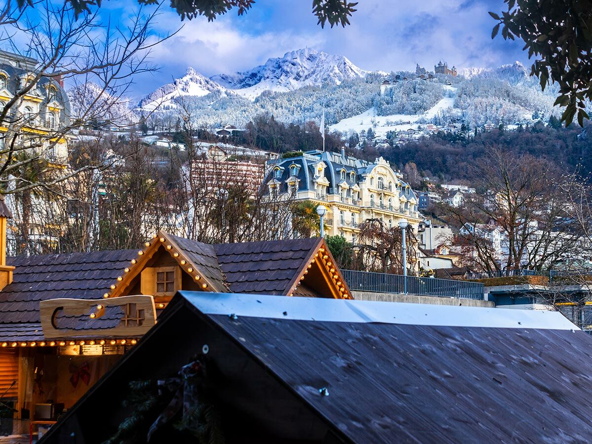 Winterliches Alpenpanorama mit schneebedeckten Bergen, Schloss, großem Hotel und beleuchteten Marktständen im Vordergrund.