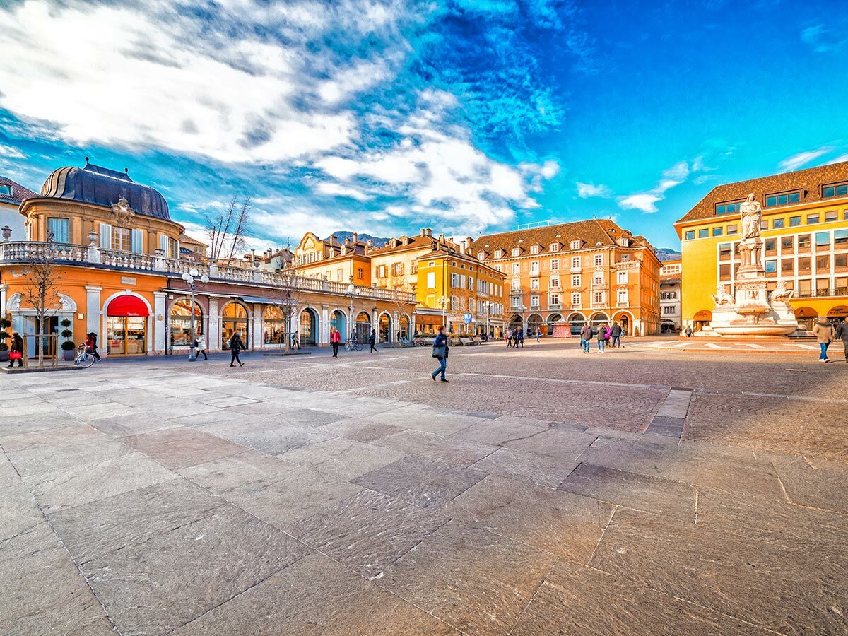 Sonnenbeschienener Waltherplatz in Bozen, Italien, mit bunten historischen Gebäuden, Menschen und einem Denkmal unter blauem Himmel.