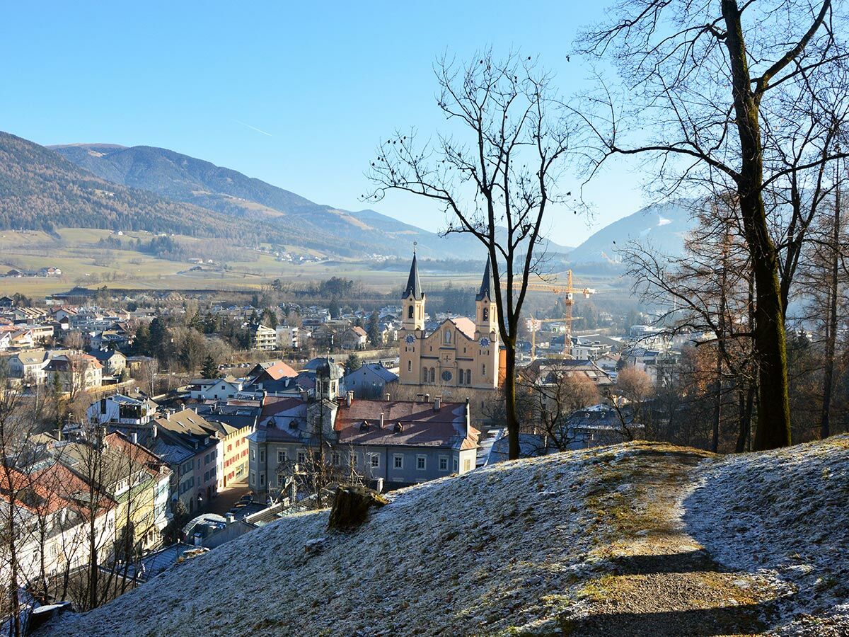 Panoramablick auf eine winterliche Bergstadt mit großer Kirche, umgeben von schneebedeckten Bergen und einem gefrorenen Wanderweg im Vordergrund.